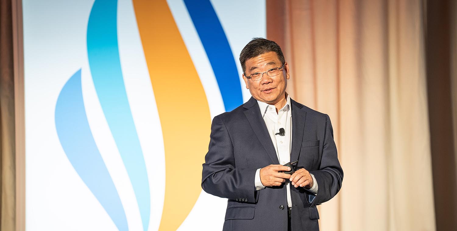 Man in suit speaking on stage with Geron logo behind.