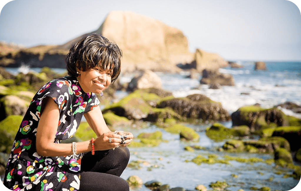 Joan smiling while sitting on a rocky, seaweed-covered shore with ocean waves in the background.