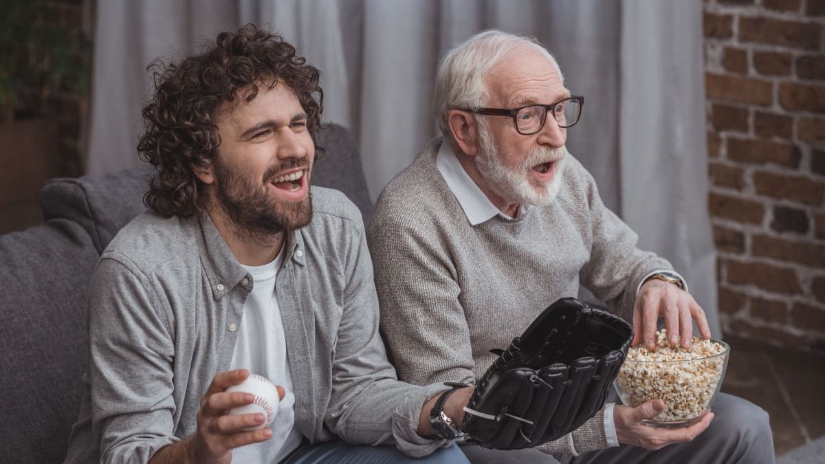 adult son and senior father cheering on baseball team and watching game at home