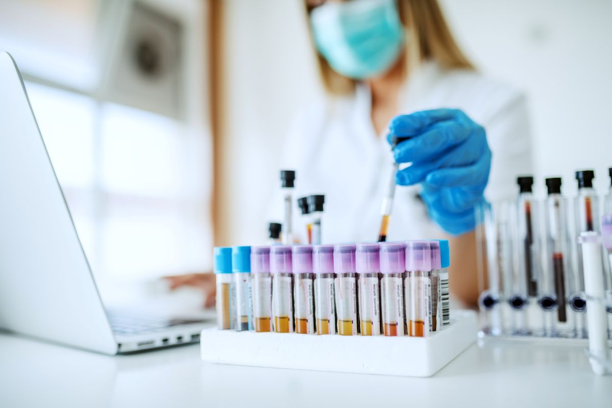Close up of lab assistant in uniform, with mask and rubber gloves holding test tube with blood sample while sitting on chair and typing on laptop.