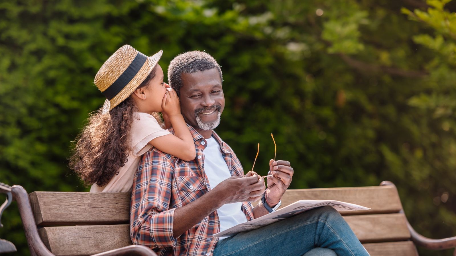 little adorable african american grandchild whispering to her smiling grandfather while sitting on bench in park